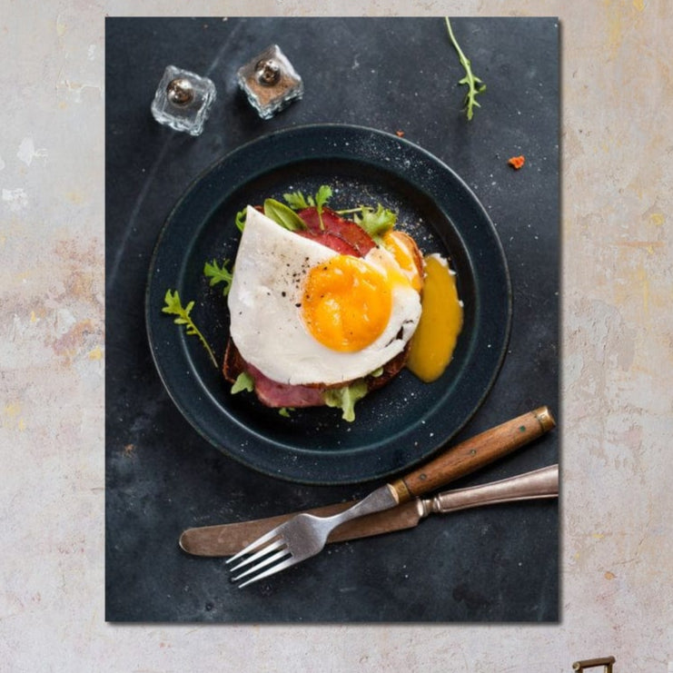 tablou canvas cu o farfurie cu un ou prajit si bacon formand un mic dejun simplu, pe o masa neagra cu tacamuri si condimente
