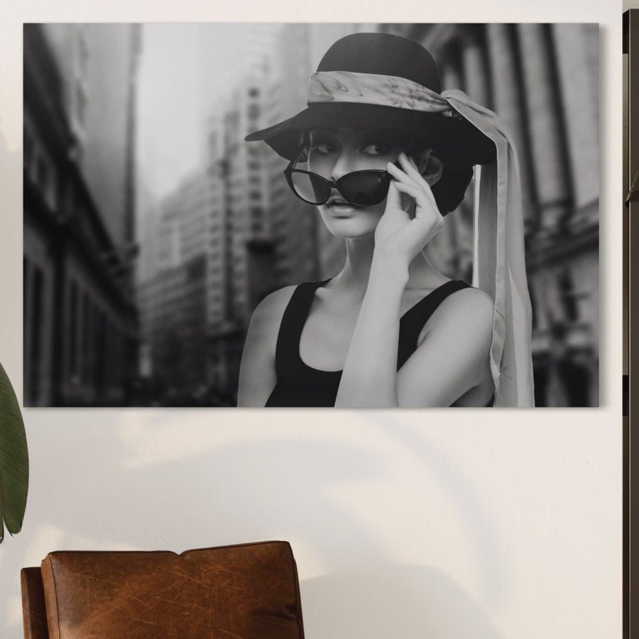 Panza LuxCanva® Tablou Canvas Premium A1356 prezintă o femeie șic cu pălărie și ochelari de soare pe clădirile orașului, în alb și negru elegant. Realizat din panza bumbac, acesta aduce stil deasupra unui scaun din piele maro și a unei plante verzi.