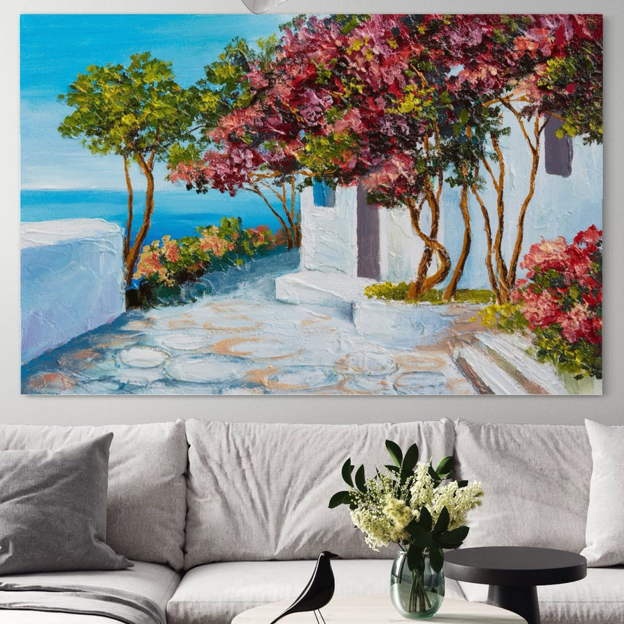 Pânza Tablou Canvas Premium LuxCanva® A1237, care prezintă un copac cu flori vibrante lângă mare și case albe, decorează peretele de deasupra unei canapele gri deschis, cu o masă rotundă cu flori albe și o figurină de pasăre neagră în față.