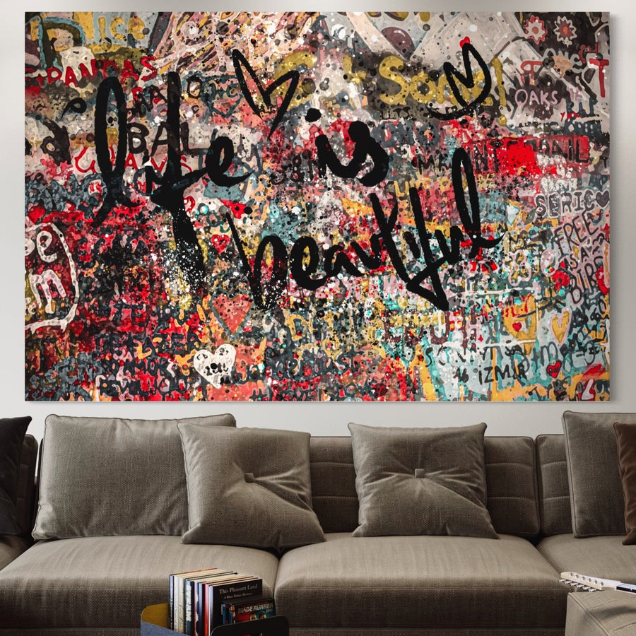 Deasupra unei canapele bej, tabloul LuxCanva® Tablou Canvas Premium LuxCanva® A1186 prezintă graffiti abstracte vibrante cu "Viața este frumoasă" în litere negre îndrăznețe, inimi și coroane - o artă murală decorativă perfectă pentru dormitoare sau camere de zi.