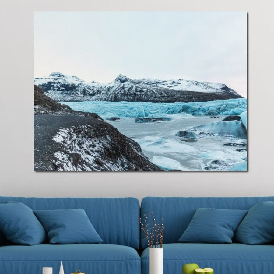 Un living modern cu o canapea albastră stă sub Tablou Canvas Premium D1042 de la LuxCanva®, Peisaj Glaciar - un peisaj înghețat cu culori vibrante, perfect pentru vibrațiile de iarnă. O mică vază albă cu ramuri decorează măsuța de cafea.