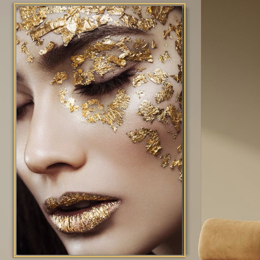 Tablou Canvas Premium LuxCanva® A1056, "Gold Eyes": portret senzual de femeie cu ochii închiși și foiță aurie artistică pe piele, în tonuri vibrante, încadrat elegant pe perete bej lângă un fotoliu crem. Brand: LuxCanva®.