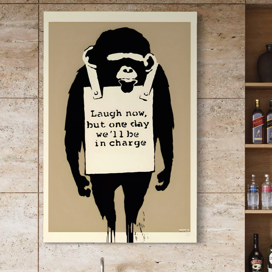 Tablou Canvas Premium LuxCanva® A1387, cu print vibrant ce prezintă o maimuță și mesajul "Laugh now, but one day we'll be in charge", expus pe un perete de piatră bej lângă rafturi cu sticle.