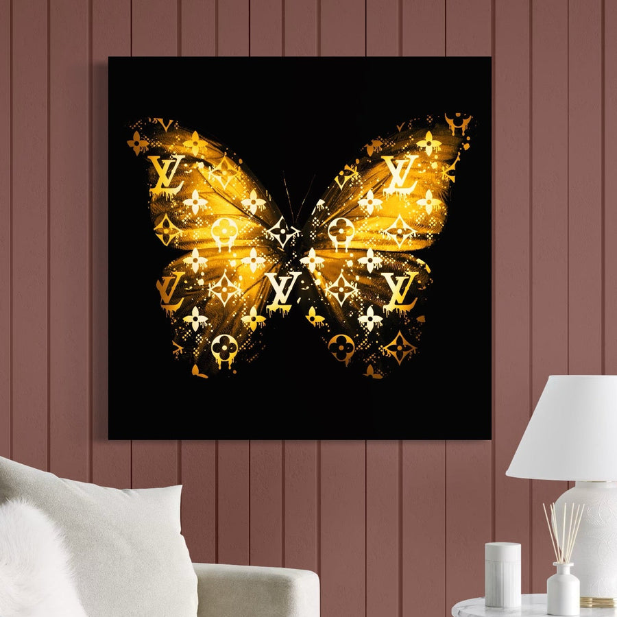 The LuxCanva® Tablou Canvas Premium B1084 prezintă Fluturele Louis Vuitton în negru și auriu pe pânză bumbac, perfect pentru livinguri moderne, deasupra unei canapele albe sau lângă o lampă albă, aducând un aer fashion și luxury încăperii.