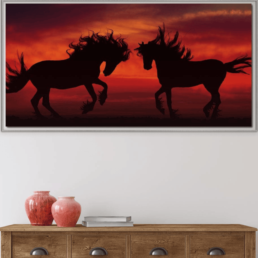 Tabloul LuxCanva® Tablou Canvas Premium "Dans în Flacăra Apusului" prezintă două siluete de cai care dansează la apus, expuse deasupra unui dulap din lemn cu două vaze roșii și cărți.