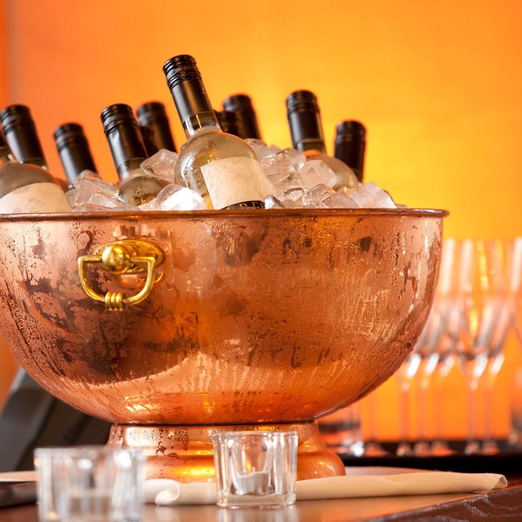 tablou canvas cu o frapiera transparenta cu sticle de vin alb si gheata, pe fundal portocaliu cu pahare si lumina calda