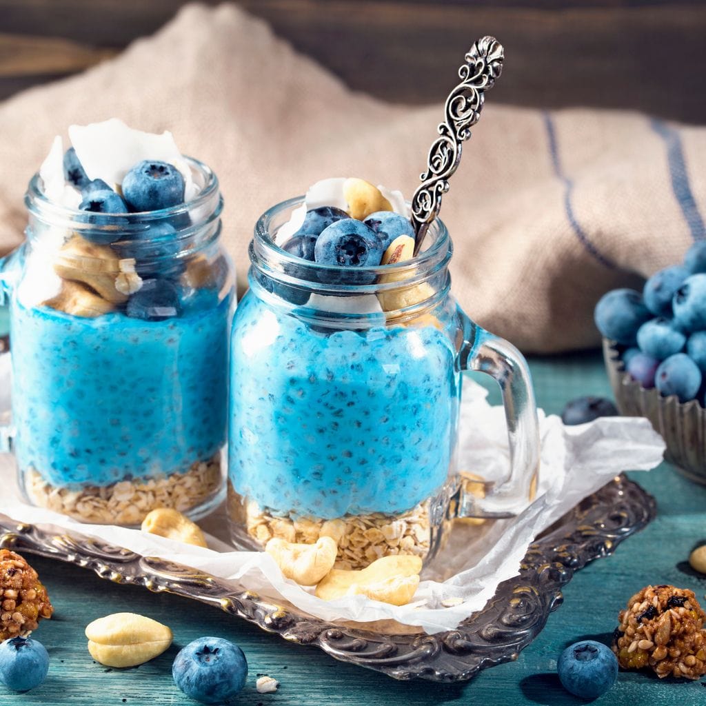 tablou canvas cu doua deserturi cu seminte de chia, afine si alune, pe o tava argintie, pe o masa albastru - turcoaz