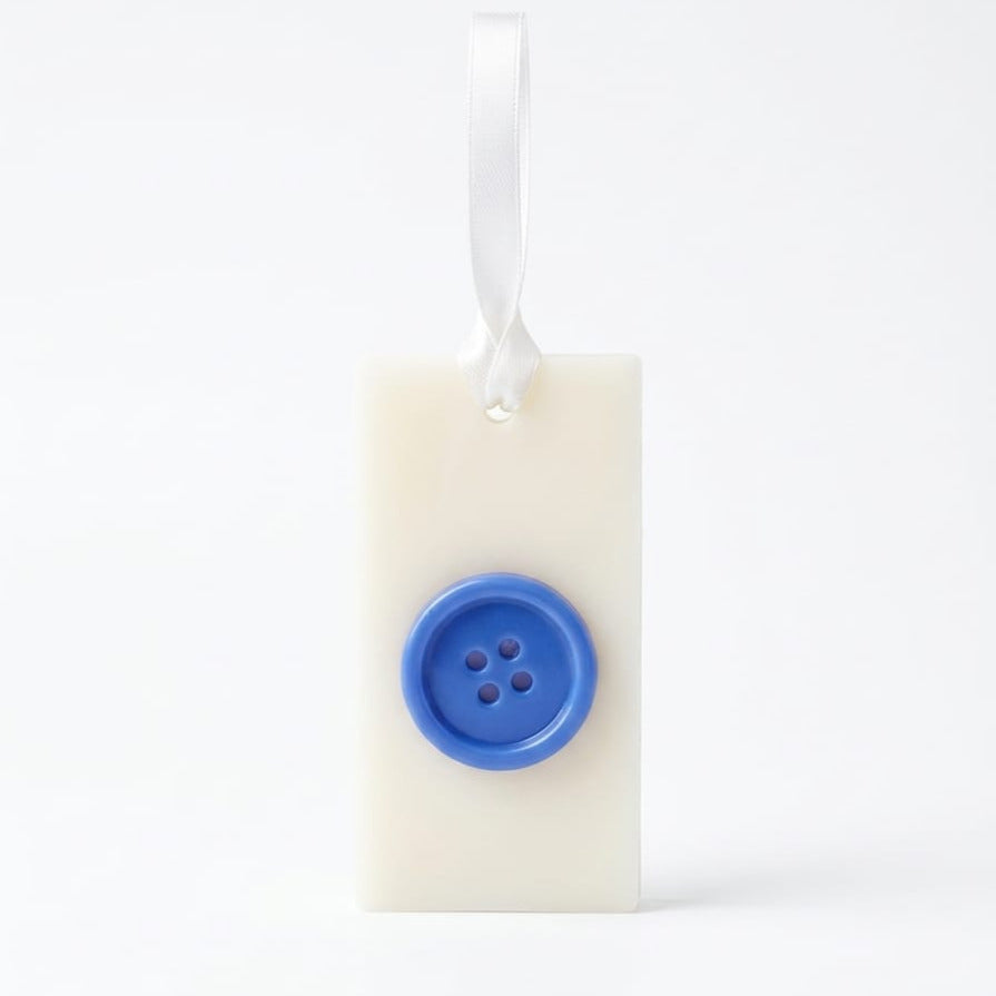 LuxCanva® Odorizant Dulap/Sertar/Pantofar GeRa Essence Gentleman este un ornament dreptunghiular alb, realizat manual, din ceară naturală (30g), cu un buton central albastru și panglică din satin, cu note de mosc alb, smirnă și tamaie.