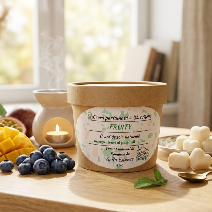 Un LuxCanva® GeRa Essence Wax Melts din soia (90 g, fructul pasiunii, mango, afine) este așezat pe o masă înconjurat de afine, felii de mango și ceară topită, în timp ce un încălzitor de ceară eliberează aburi în lumina soarelui prin intermediul unei ferestre.